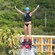 A woman on a flyboard holds her hands out while being propelled high in the air by a long hose connected to a jet ski